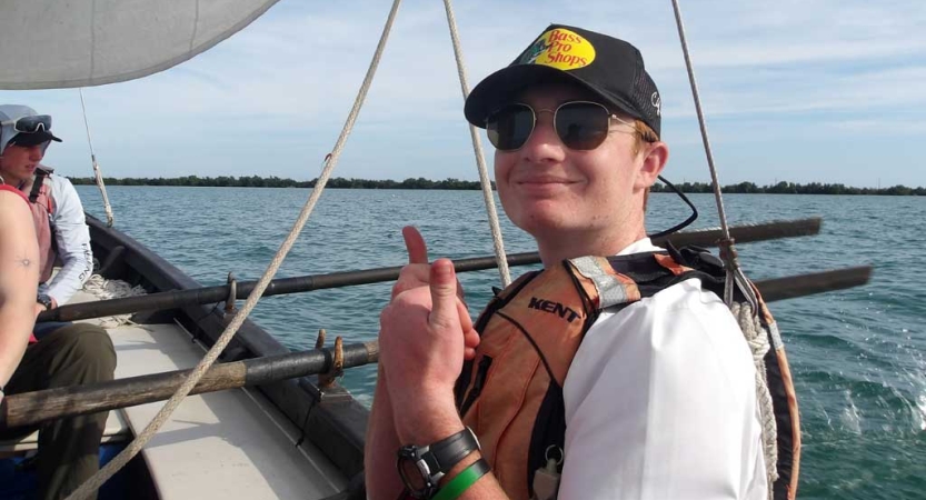 a person wearing a life jacket smiles and gives two thumbs up while sitting in a sailboat. 